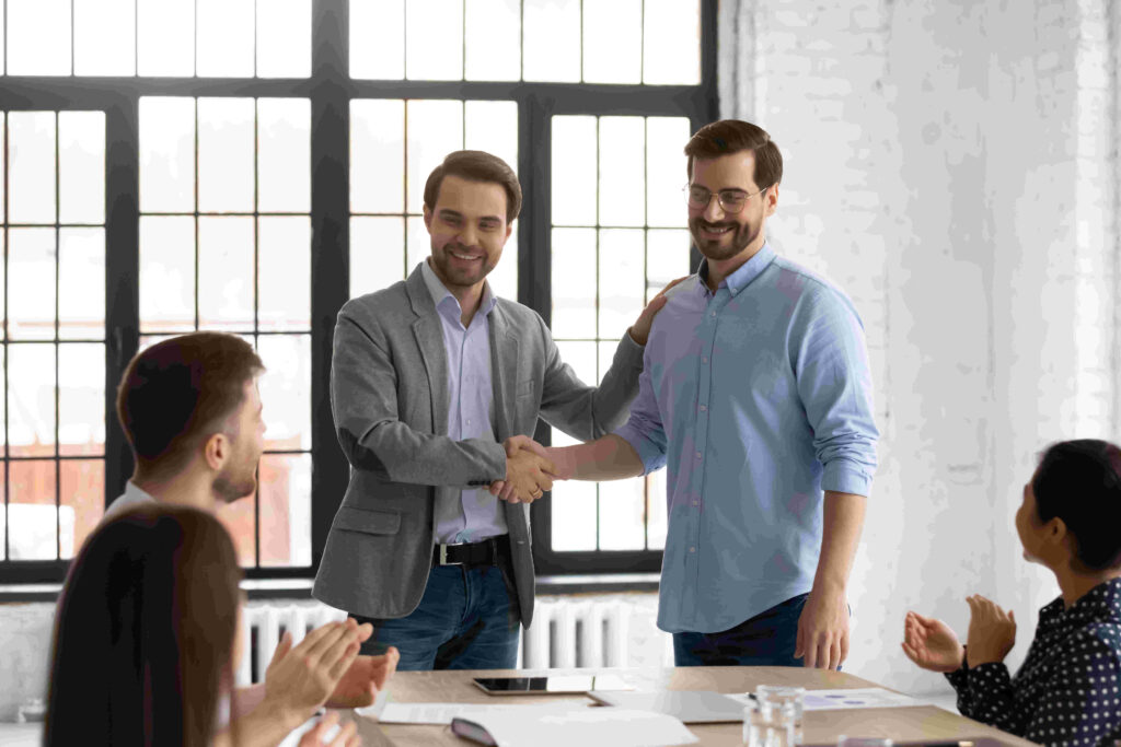 Two business men shaking hands as one congratulates the other and a group clap in the foreground